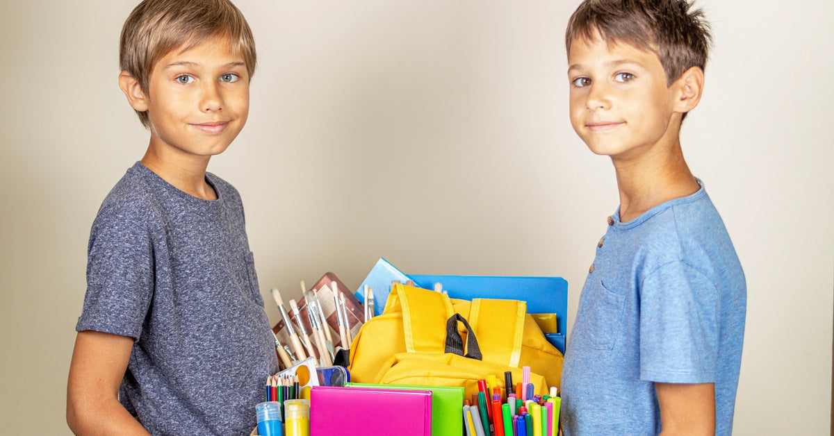 Two smiling young boys hold a box between them. The box contains colorful pencils, markers, backpacks, and folders.