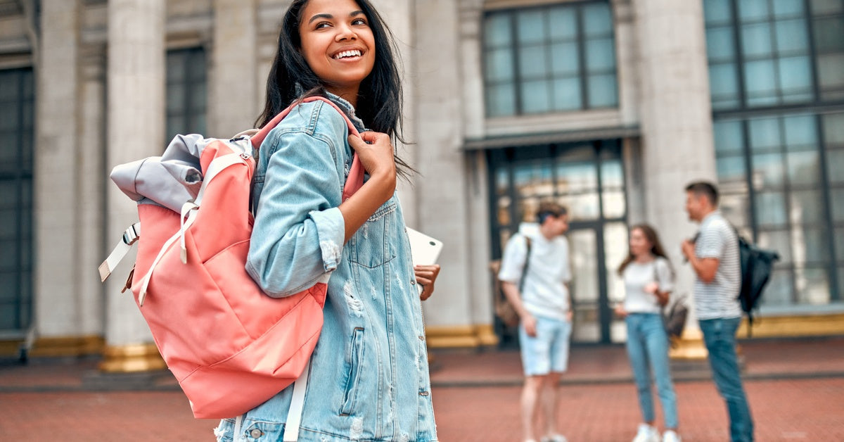 A smiling girl with a pink backpack and laptop looks over her shoulder. There are three students in the background.
