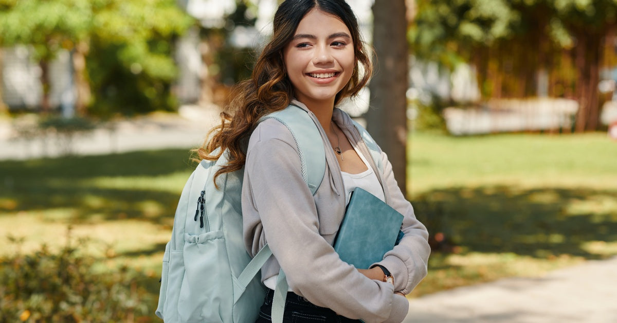 A smiling young girl carrying a notebook and backpack stands on a sidewalk and looks over her shoulder.