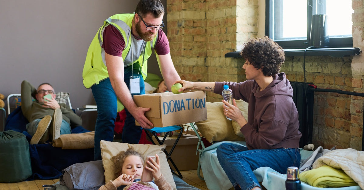 A man in a safety vest bending down and offering a donation box to a seated woman. She's holding a water bottle and an apple.