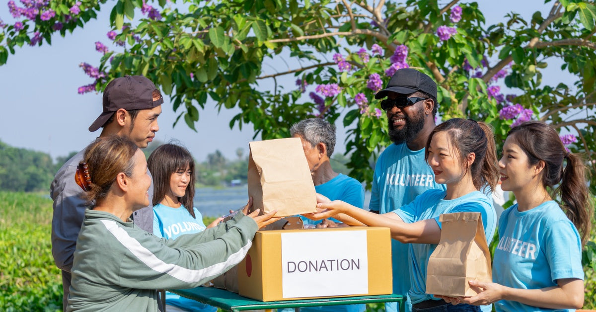 Smiling male and female volunteers in blue shirts are distributing bagged donations near a large tree with purple flowers.