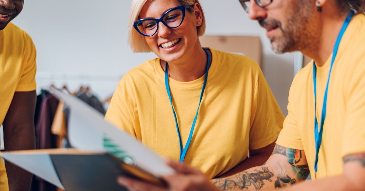 Two smiling people in yellow shirts are examining papers. Stacked boxes and a rack of clothing are behind them.