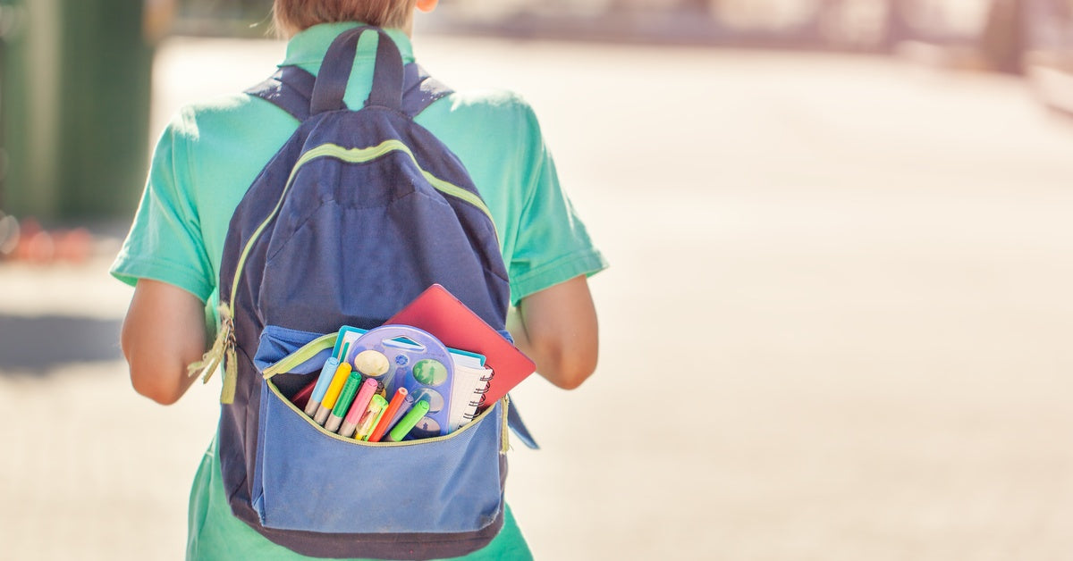 A schoolboy wearing a blue backpack with an open front pocket full of notebooks, folders, and markers.