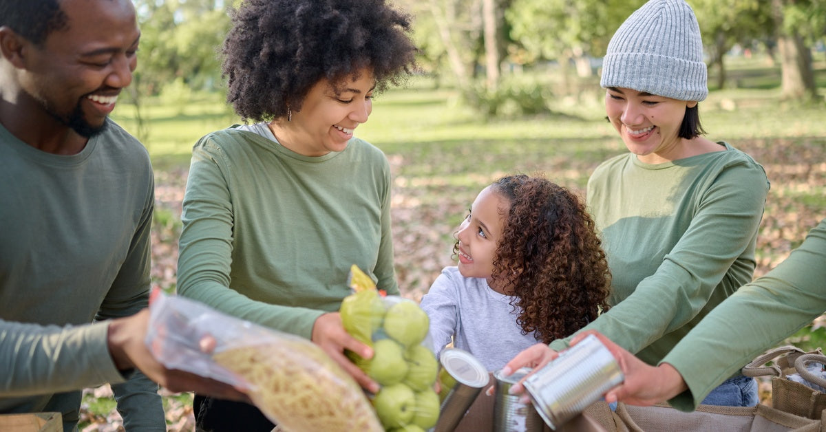 Smiling adults and a little girl standing in a tree-filled park and placing food donations into a cardboard box.