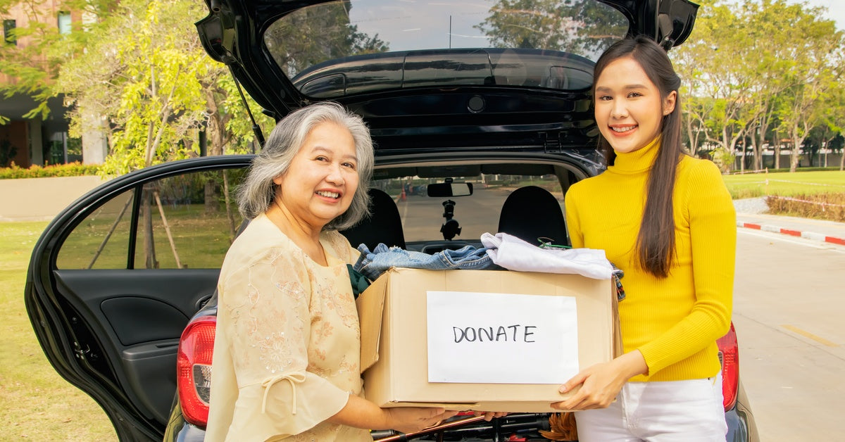 An older woman and a younger woman standing by an open car trunk, holding a cardboard box labeled '"donate."
