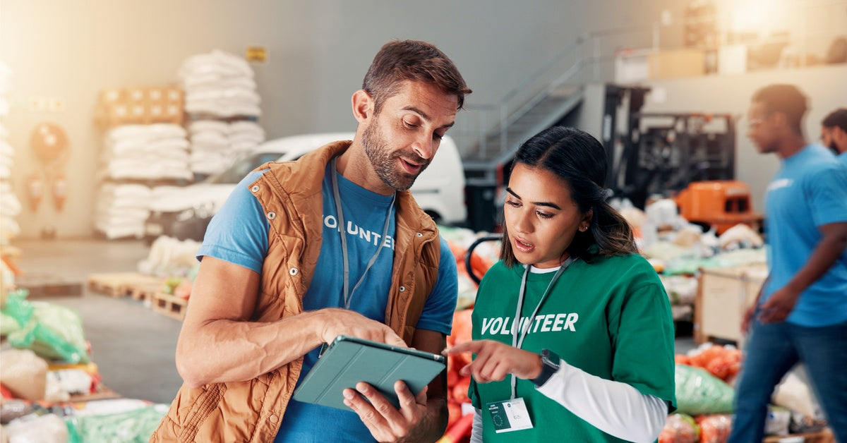 Two people with "volunteer" written on their shirts consulting a tablet inside a room filled with bagged donations.