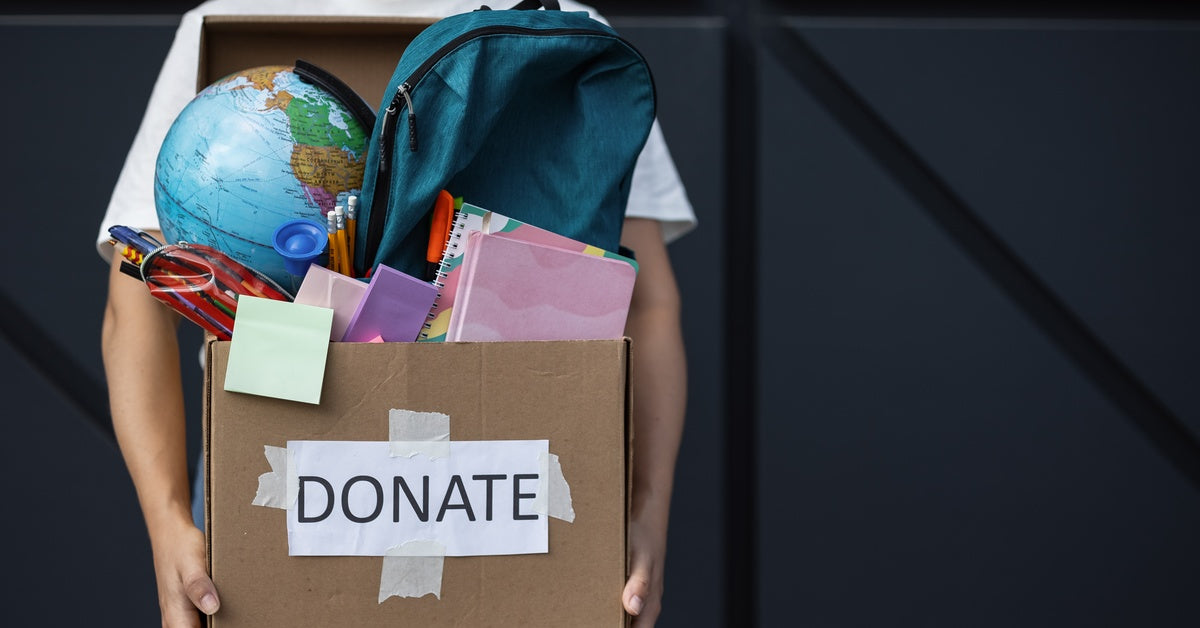 A close-up of a person holding a box filled with school supplies, with a sign reading "donate" on the front.