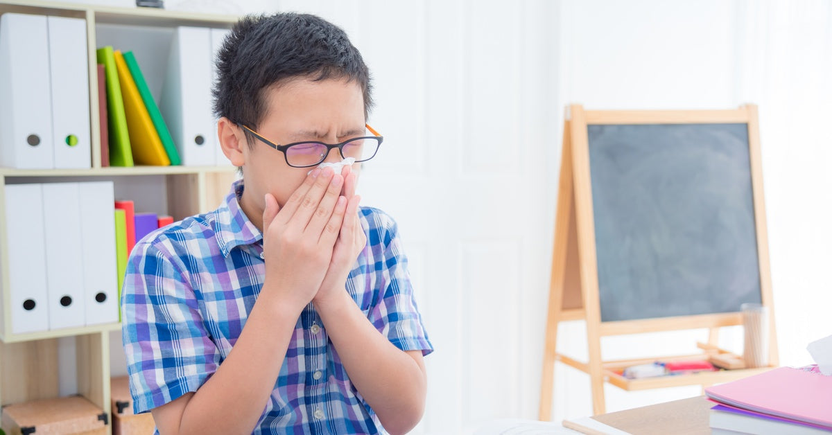 A young boy wearing a blue flannel shirt and glasses sits at a desk with a textbook open as he blows his nose.