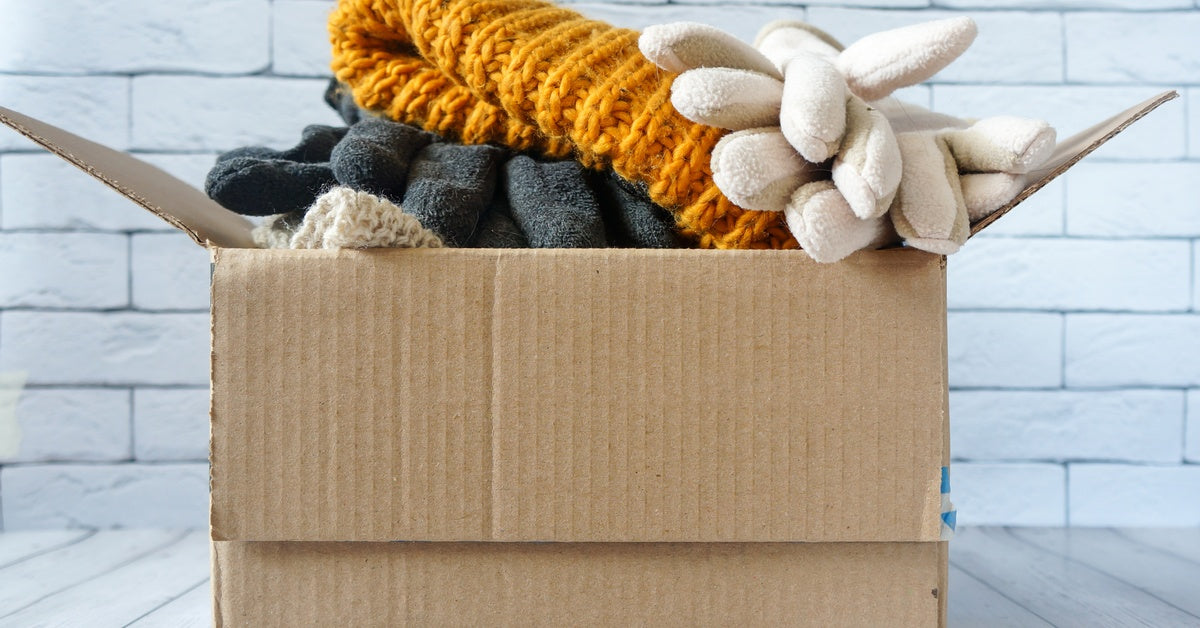A cardboard box on a wooden surface holds a yellow knit beanie and gloves, set against a white brick wall.