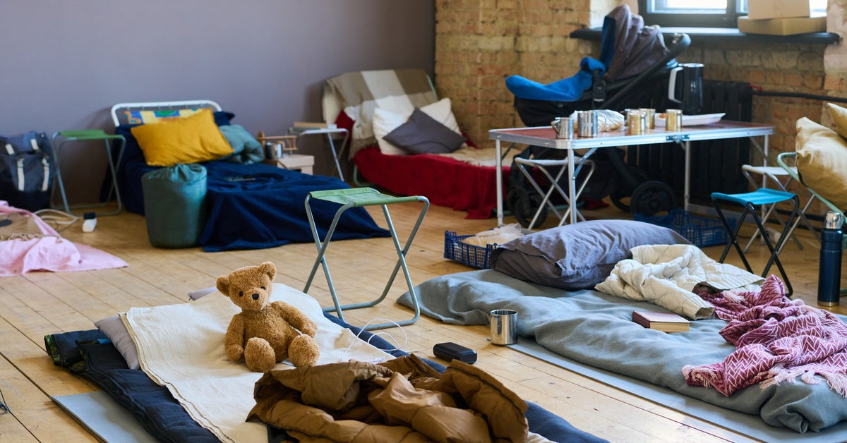 A spacious room features several sleeping arrangements on a wooden floor, next to a bright window along a brick wall.