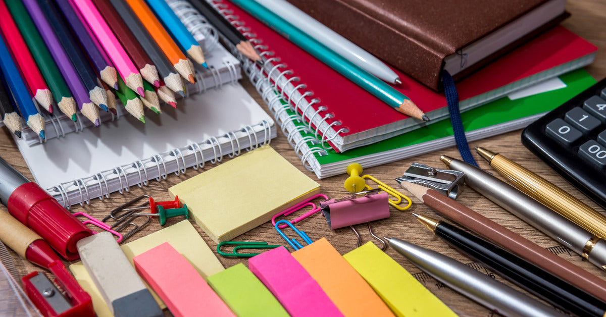 An array of colorful back-to-school supplies sit on a wooden table. There are colored pencils, erasers, notebooks, and pens.