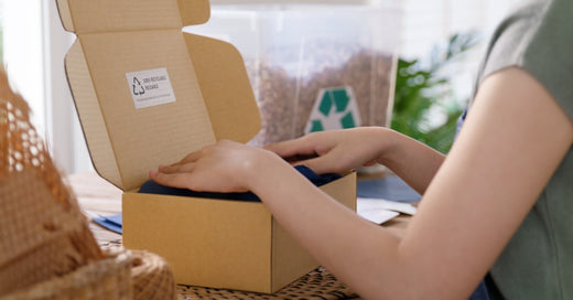 A woman's hands making a box with a recycling label on the inside. There is a clear box of recyclable packing peanuts.