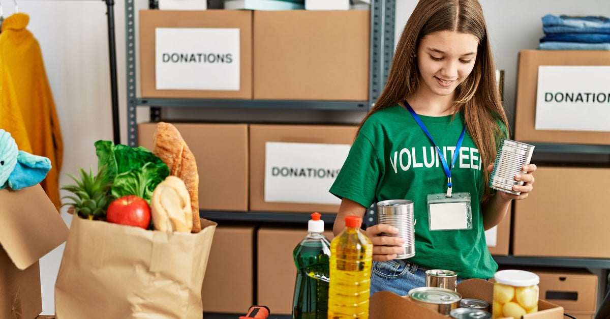 A young teen girl in a green volunteer shirt looking through a box of canned goods next to a box of children's toys.