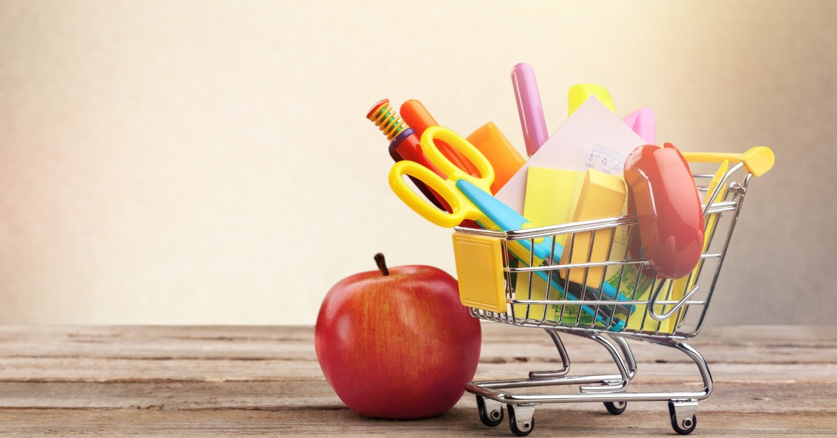 A miniature shopping cart filled with school supplies, including scissors and pens, is placed next to an apple.
