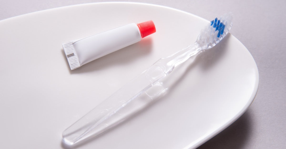 A close-up of a transparent toothbrush and a small tube of toothpaste resting on a white dish atop a gray surface.
