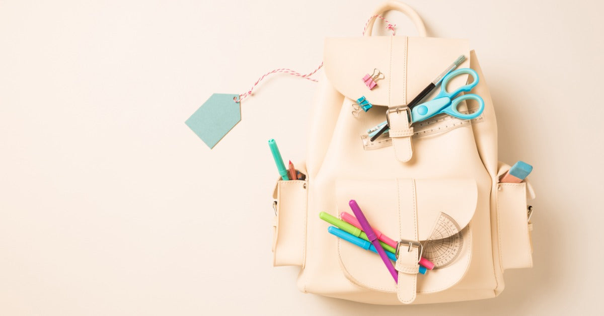 A white backpack contains various school supplies such as scissors, an eraser, and pens in different colors.