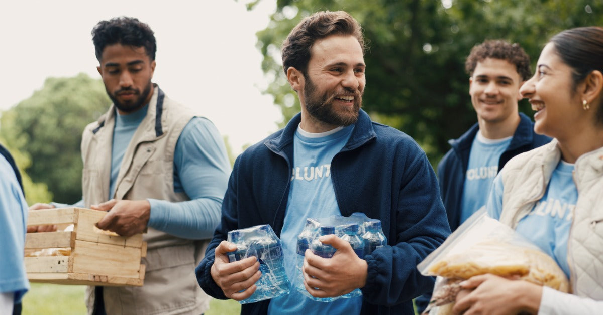 A man carrying two bundles of packaged water bottles walks with other young adults in an outdoor area.