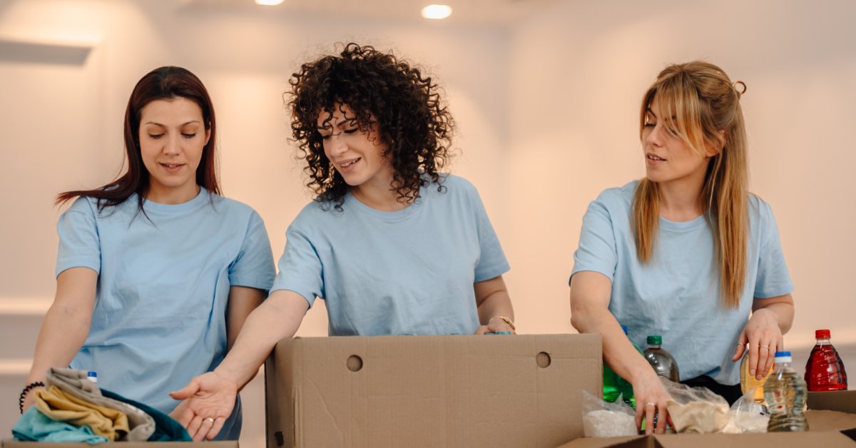 Three Caucasian women stand at a table organizing a variety of supplies as they pack them in cardboard boxes.