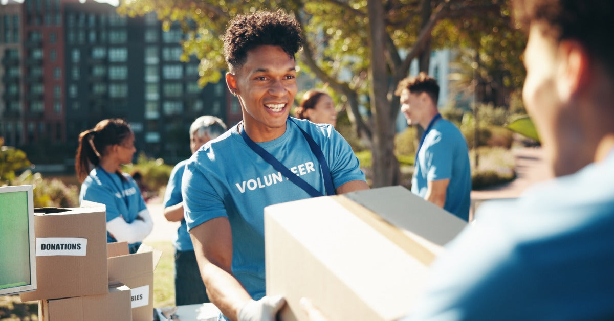 Two young men carry a cardboard box at a community park while several boxes labeled “Donations” sit stacked on a table.