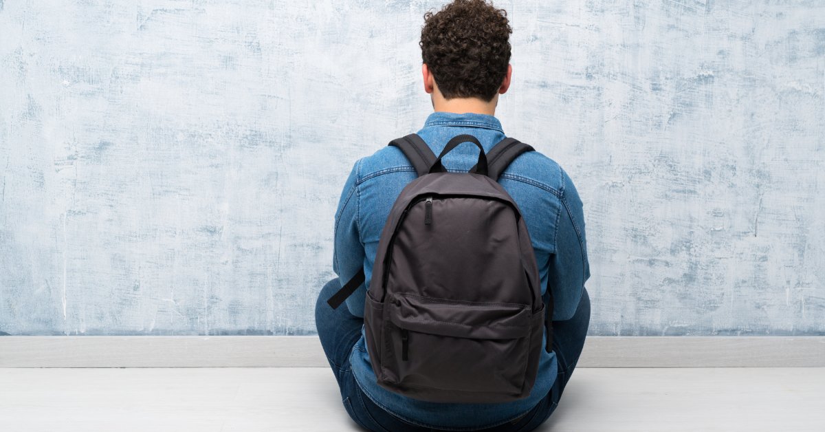 A person with curly hair sitting on the floor in front of a blue wall, wearing a black backpack and denim shirt.