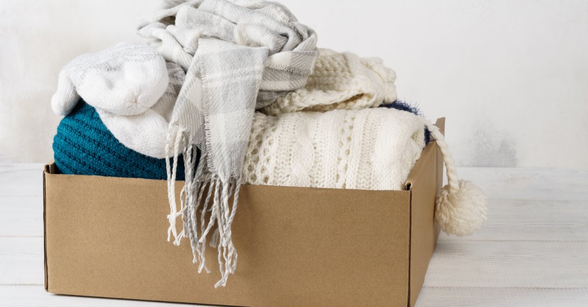 A cardboard box filled with folded knit sweaters, a white scarf, and winter hats on a light wooden table.