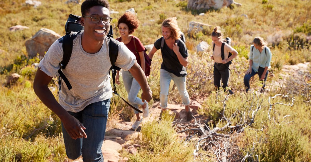 Smiling young adults with backpacks walking single file on an uphill path surrounded by rocks and low bushes.