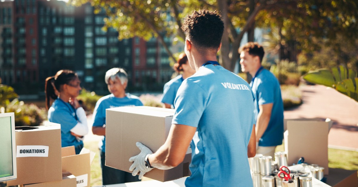 A volunteer in a blue shirt and gloves carries a cardboard box. He approaches a group of other helpers.