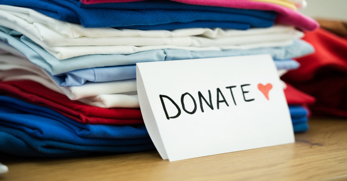 A close-up of a stack of clothes in various colors on a wooden surface, with a folded paper sign that reads "donate."