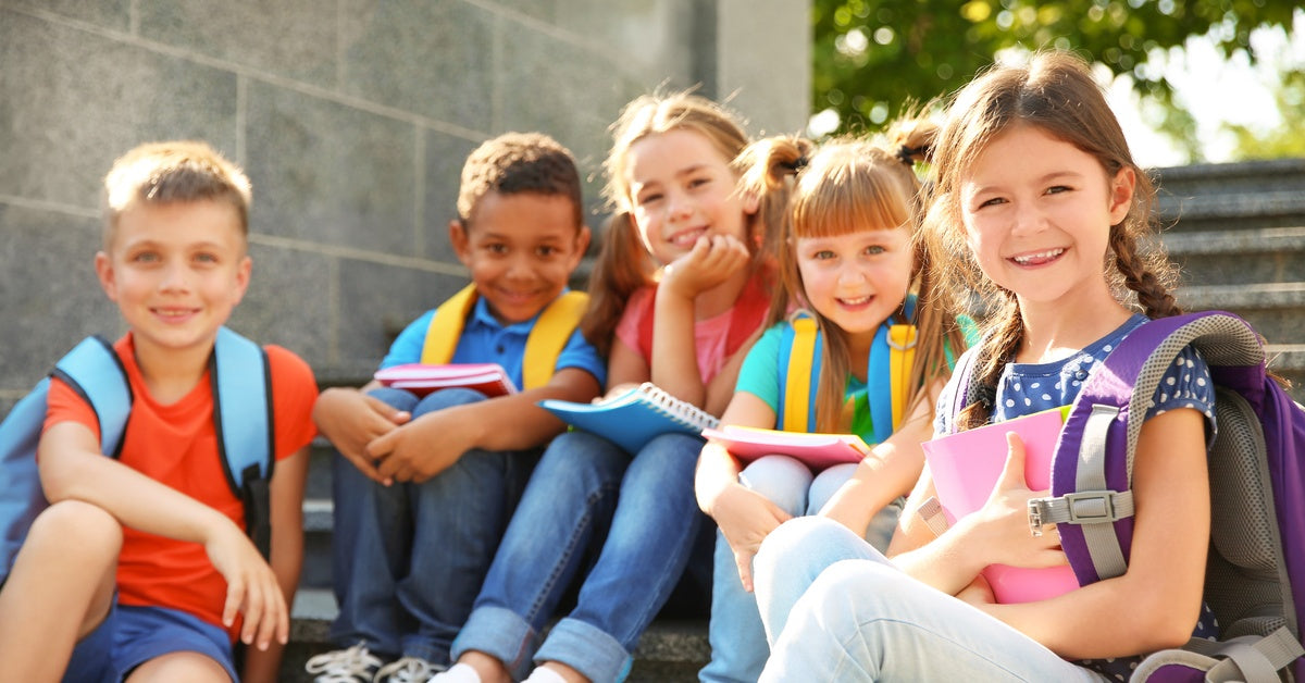 A group of three girls and two boys smiling as they sit outside on stairs near a brick wall on a sunny day.