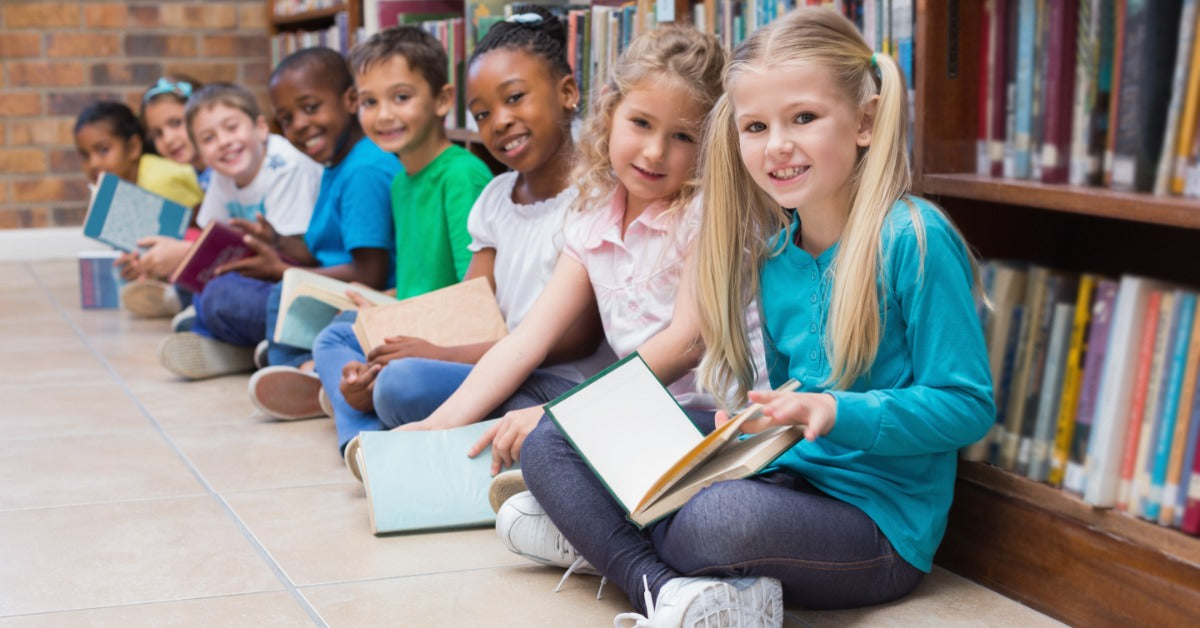 Eight children sit cross-legged on the floor in front of a bookshelf, each smiling and holding a different book.