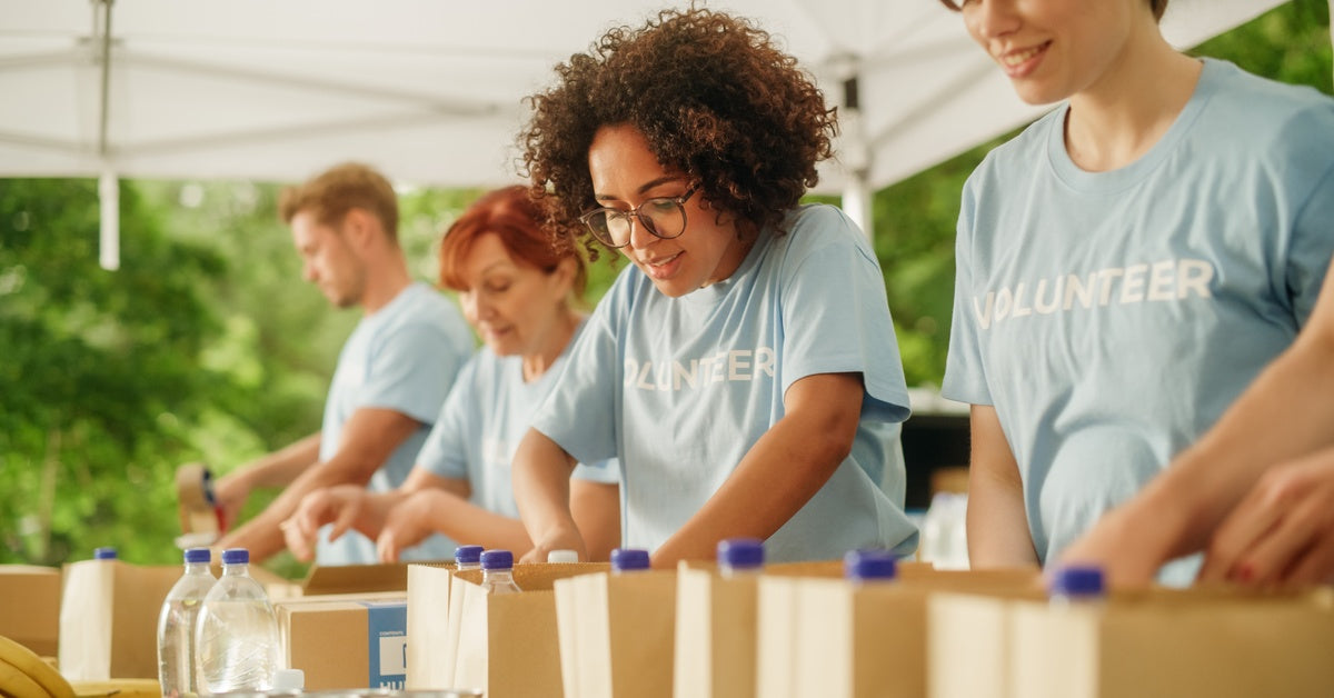 Several people wearing blue shirts that say "volunteer" stand in a line under a white tent, packing brown bags.