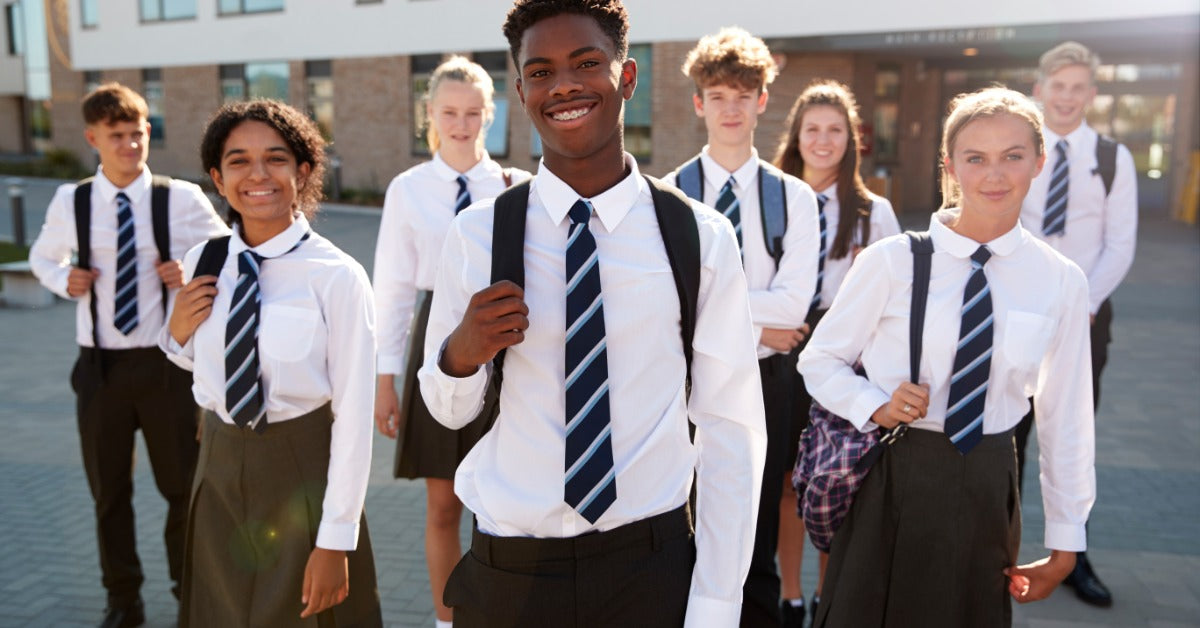 A group of diverse high school students in matching uniforms stands outside of a building on a sunny day, smiling.