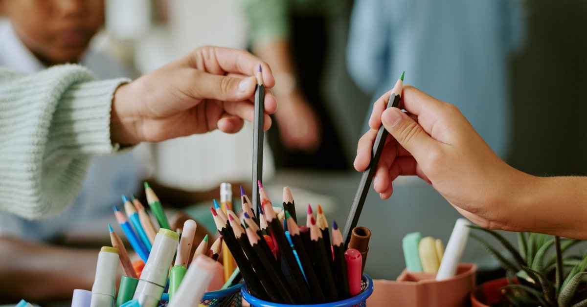 A close-up of two kids reaching for colored pencils from a container. Two people are visible in the background.