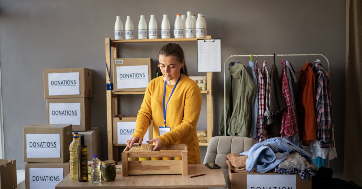 A person standing beside shelves and cardboard boxes filled with food and supplies in a donation storage area.