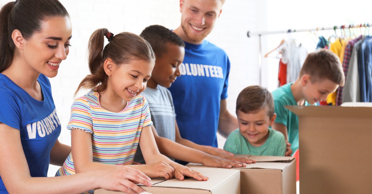 Adult volunteers and children sort through cardboard boxes and toys in a bright room with clothing racks.