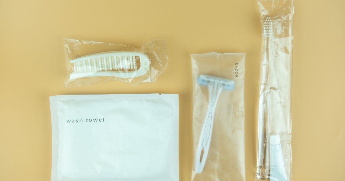 Packaged travel toiletries arranged on a beige surface, including a comb, razor, toothbrush, and wash towel.