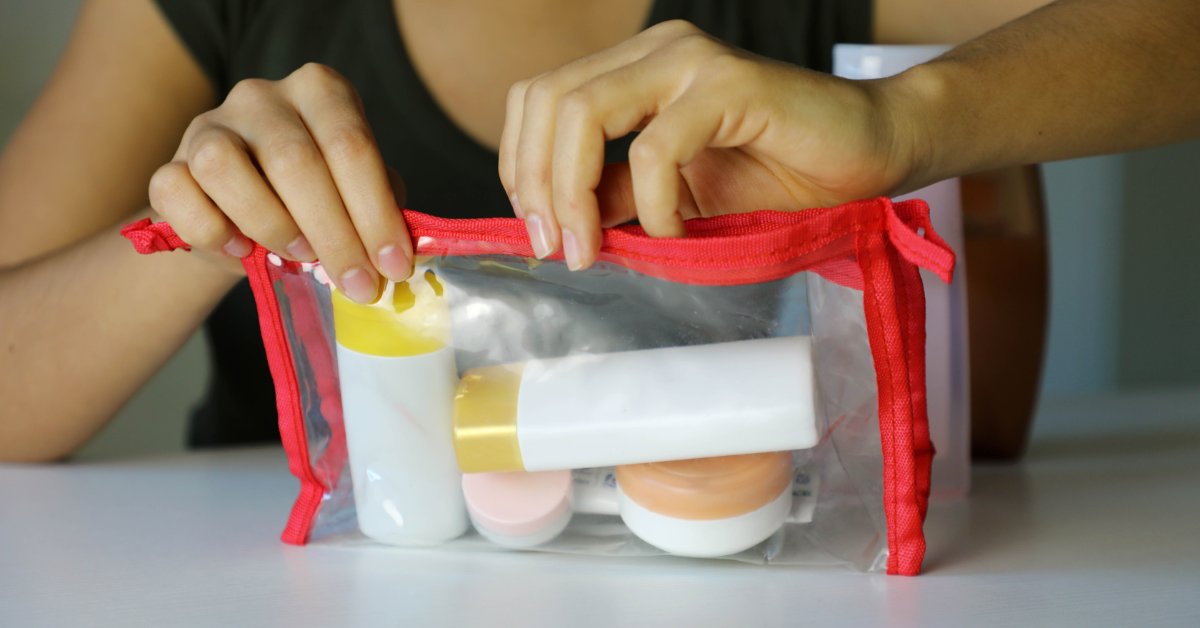 A close-up of a person closing a clear toiletry bag filled with small bottles and travel items on a white surface.