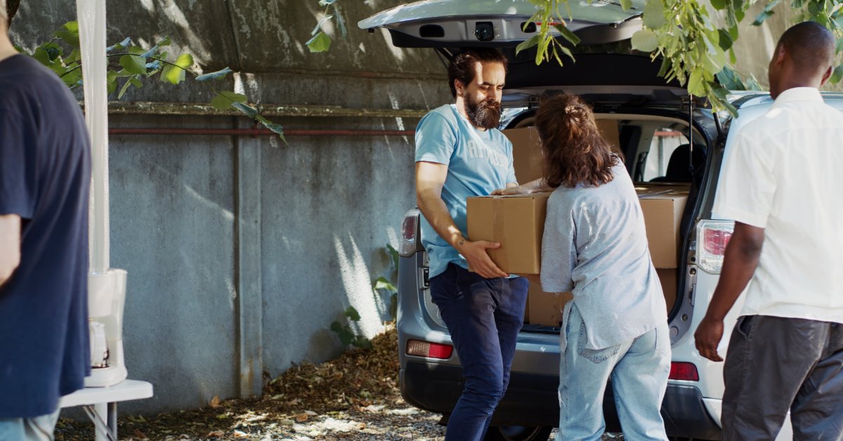A close-up of a volunteer handing a donation box to two people beside a car with its trunk open under a tree.