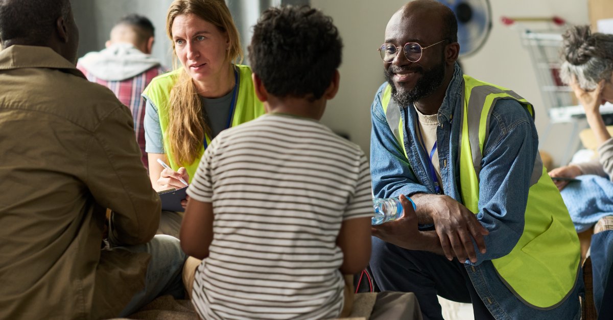 A woman with a clipboard and a man wearing a yellow safety vest are talking with an adult and a child seated indoors.